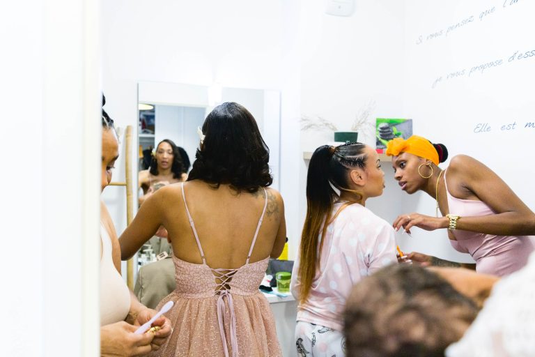 Un groupe de femmes se prépare dans une salle de bain lumineuse. Une femme en robe rose se tient devant un miroir. Une autre ajuste le maquillage d'une femme portant un peignoir à pois. Une quatrième est partiellement visible, reflétée dans le miroir, avec "Mariage Audenge" élégamment écrit sur un mur.