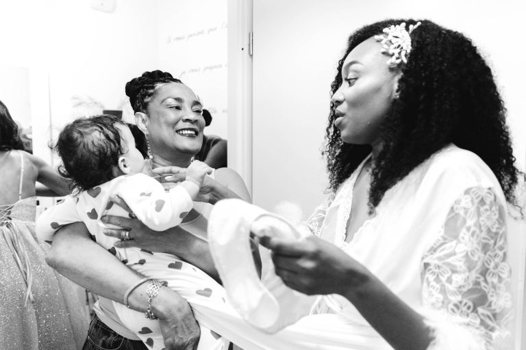 Image en noir et blanc d'une femme souriante tenant un bébé vêtu de vêtements à motifs de cœurs, tandis qu'une autre femme en robe blanche, aux cheveux bouclés et avec un accessoire pour cheveux, interagit avec eux. Les deux femmes semblent être dans une cabine d'essayage. L'ambiance semble joyeuse et intime, rappelant les célébrations du Mariage Audenge.