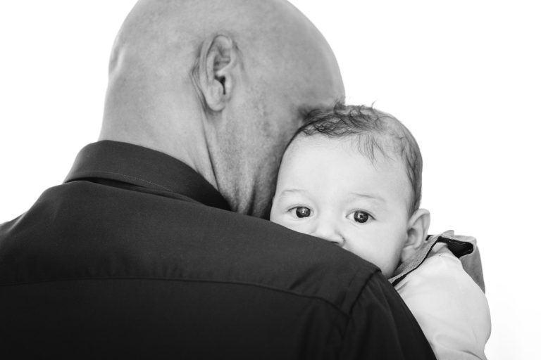 Gros plan en noir et blanc d'un adulte tenant un bébé dans ses bras. Le dos de l'adulte est tourné vers l'objectif, tandis que le bébé regarde droit devant lui, la tête appuyée sur l'épaule. Les yeux écarquillés et l'expression calme, le bébé regarde directement l'objectif, capturant un moment touchant évoquant une célébration de Mariage Audenge.