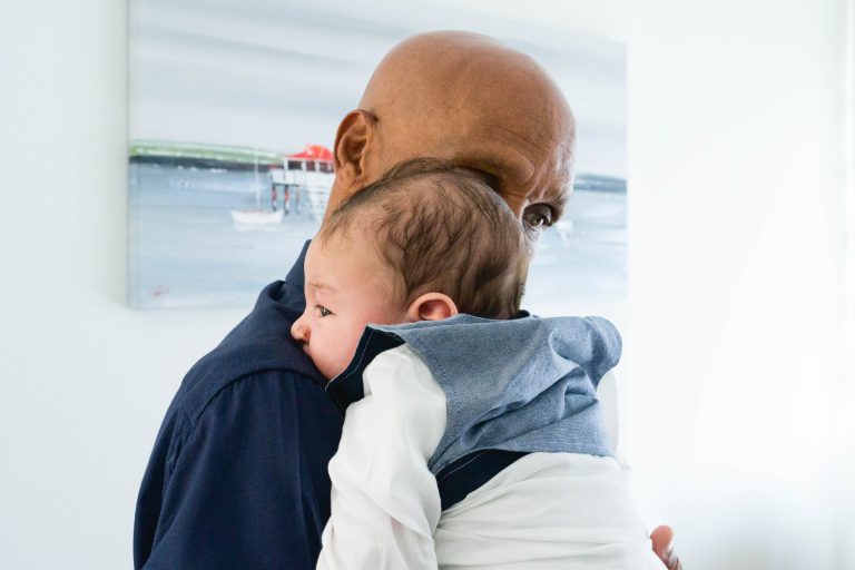 Un homme chauve, vêtu d'une chemise sombre, tient un bébé contre son épaule. Le bébé, vêtu d'une combinaison blanche et d'un gilet gris clair, s'appuie confortablement sur l'épaule de l'homme. Dans l'arrière-plan flou, on voit un tableau représentant une scène côtière avec des bateaux et un bâtiment au toit rouge rappelant Mariage Audenge.