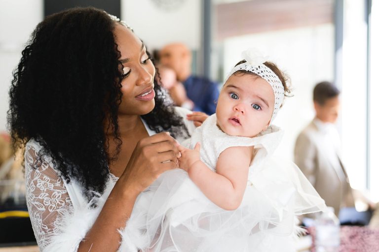 Une femme aux cheveux longs bouclés, vêtue d'une tenue en dentelle blanche, tient dans ses bras un bébé vêtu d'une robe blanche avec un bandeau assorti. La femme regarde affectueusement le bébé dans ses bras. L'arrière-plan du Mariage Audenge est légèrement flou avec quelques personnes visibles au loin.