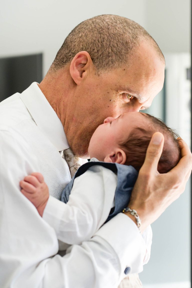 Un homme chauve en chemise blanche et cravate berce affectueusement un bébé dans ses bras, capturant peut-être un moment précieux du mariage Audenge. Il tient doucement la tête du bébé et se penche pour toucher le nez du nourrisson, qui est vêtu d'une chemise blanche et d'un gilet bleu. L'arrière-plan légèrement flou attire l'attention sur leur tendre connexion.