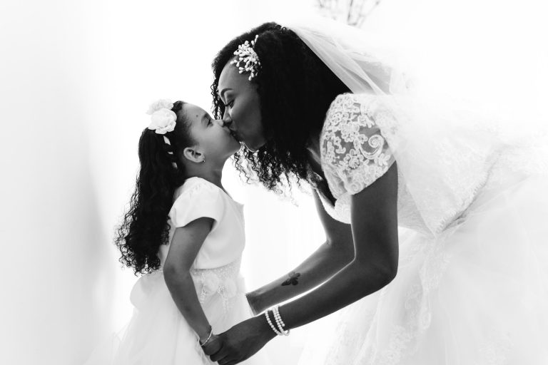 Une mariée en robe de mariée en dentelle et voile se penche pour embrasser une jeune fille en robe blanche et coiffe fleurie. Toutes deux ont les cheveux bouclés et le moment est tendre et affectueux. La photo en noir et blanc de ce Mariage Audenge capture parfaitement le lien émotionnel qui existe entre elles.