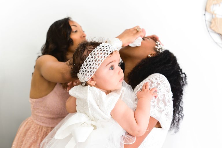 Une femme en robe rose poudré donne un biberon à une femme en robe de dentelle blanche, qui regarde vers le haut. La femme en blanc tient un bébé habillé de blanc avec un bandeau sur la tête. Le bébé, aux cheveux bouclés, regarde sur le côté avec une expression curieuse, sur fond simple de Mariage Audenge.