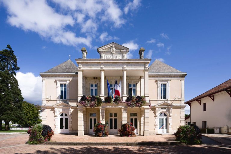 L'image montre un bâtiment historique de l'hôtel de ville français avec une façade symétrique et néoclassique. Le bâtiment comporte deux étages, de grandes fenêtres et une horloge au-dessus de l'entrée principale. Des drapeaux français et des compositions florales ornent le balcon, idéal pour le Mariage Audenge. Une cour pavée se trouve devant sous un ciel bleu clair.