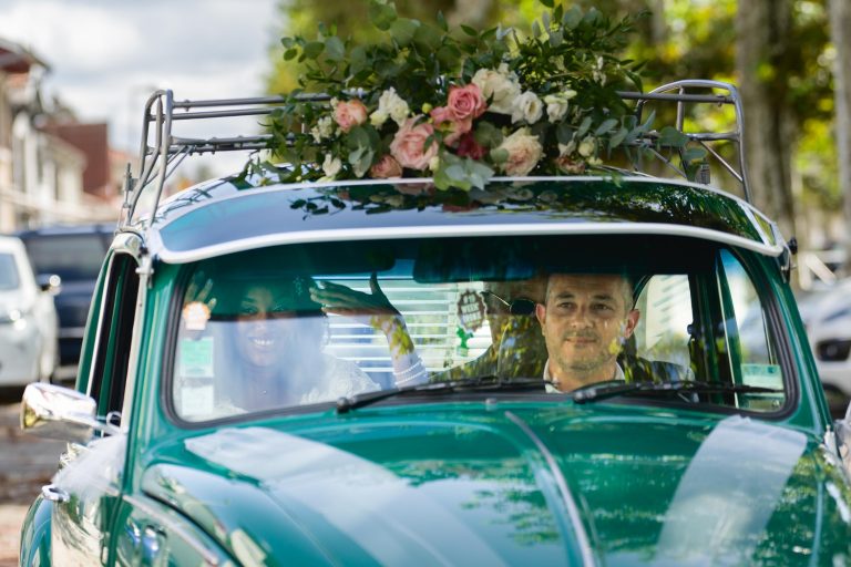 Un couple souriant à bord d'une voiture verte vintage ornée d'une grande composition florale sur le toit. L'homme conduit tandis que la femme, partiellement visible, salue depuis le siège passager. Des arbres et des bâtiments sont visibles en arrière-plan, suggérant un cadre extérieur animé pendant la célébration de leur Mariage Audenge.