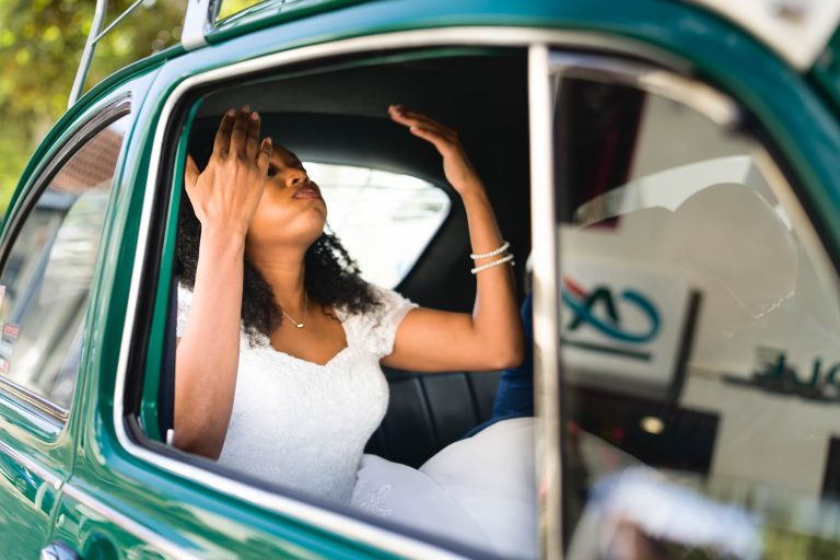 Une mariée en robe de mariée blanche aux détails en dentelle ajuste son voile tout en étant assise à l'arrière d'une voiture verte vintage, rappelant un Mariage Audenge intemporel. Elle a les cheveux bouclés et porte un bracelet. La fenêtre ouverte de la voiture révèle un arrière-plan flou avec une signalisation moderne et de la verdure.