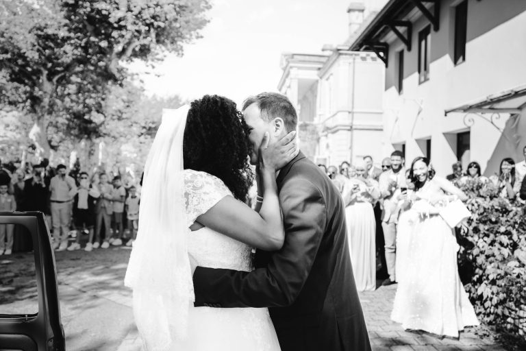 Un couple de jeunes mariés s'embrasse et s'embrasse à l'extérieur, entouré d'amis et de membres de la famille qui applaudissent à leur mariage Audenge. La mariée porte une robe en dentelle blanche et un voile, tandis que le marié porte un costume sombre. L'arrière-plan présente des arbres et un bâtiment avec des gens de chaque côté, célébrant l'occasion. L'image est en noir et blanc.