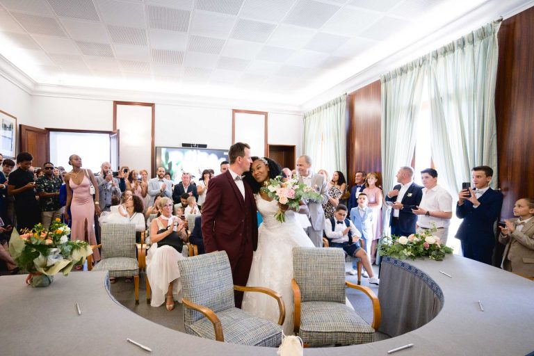 Une mariée en robe blanche et voile tient un bouquet de fleurs tandis qu'elle embrasse le marié en costume marron. Ils se tiennent dans une pièce avec de grandes fenêtres et des rideaux, entourés d'invités capturant l'instant du Mariage Audenge. Des compositions florales décoratives bordent la table incurvée où certains invités sont assis tandis que d'autres sont debout.