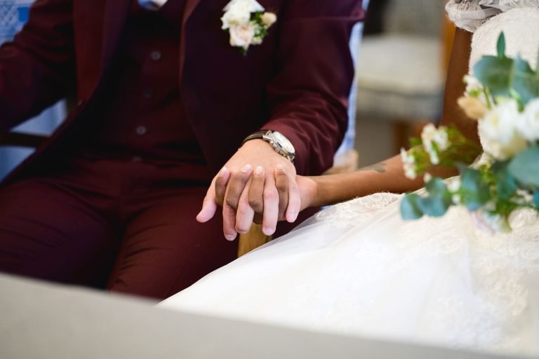 Gros plan d'une mariée et d'un marié se tenant la main lors de leur cérémonie de Mariage Audenge. La robe en dentelle blanche et le bouquet de fleurs de la mariée sont partiellement visibles, tandis que le marié porte un costume marron et une cravate, avec une boutonnière sur son revers. L'accent est mis sur leurs mains, symbolisant leur union.