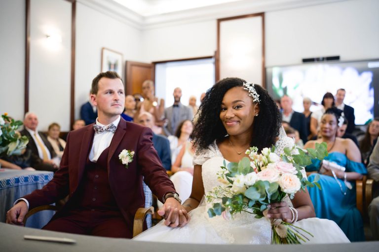 Une mariée et un marié sont assis côte à côte, se tenant la main lors de leur mariage Audenge. Le marié porte un costume marron et un nœud papillon, tandis que la mariée éblouit dans une robe blanche, tenant un bouquet de fleurs. Ils sont entourés d'invités assis et debout qui observent attentivement la cérémonie.