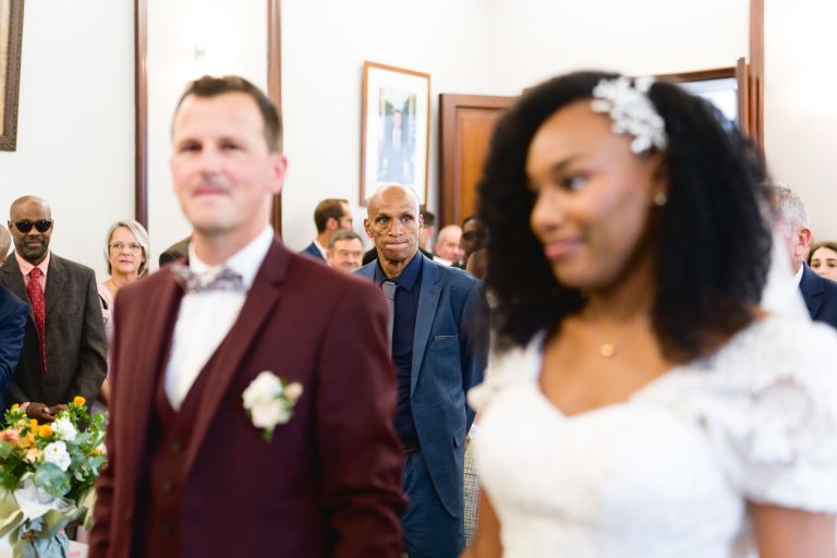 Au premier plan, un couple de mariés se tient en évidence, le marié portant un costume marron et la mariée une robe blanche aux détails floraux. À l'arrière-plan, un homme chauve en costume bleu se tient au centre, regardant directement vers l'objectif, entouré des invités du mariage de Mariage Audenge.