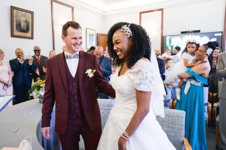 Un couple de mariés souriants se tient devant une salle remplie d'invités qui applaudissent à Audenge. La mariée porte une robe en dentelle blanche avec un accessoire de cheveux fleuri, et le marié enfile un costume marron avec un nœud papillon. Les invités, dont des femmes tenant des enfants, sont assis et debout à l'arrière-plan, célébrant leur mariage.