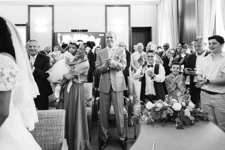 Une photo en noir et blanc capture une cérémonie intime du Mariage Audenge. Les invités sont debout et applaudissent, regardant la mariée qui est positionnée dos à la caméra. Une femme en robe tient un enfant en bas âge et une composition florale décore une table au premier plan. La salle est bien éclairée grâce à de grandes fenêtres.