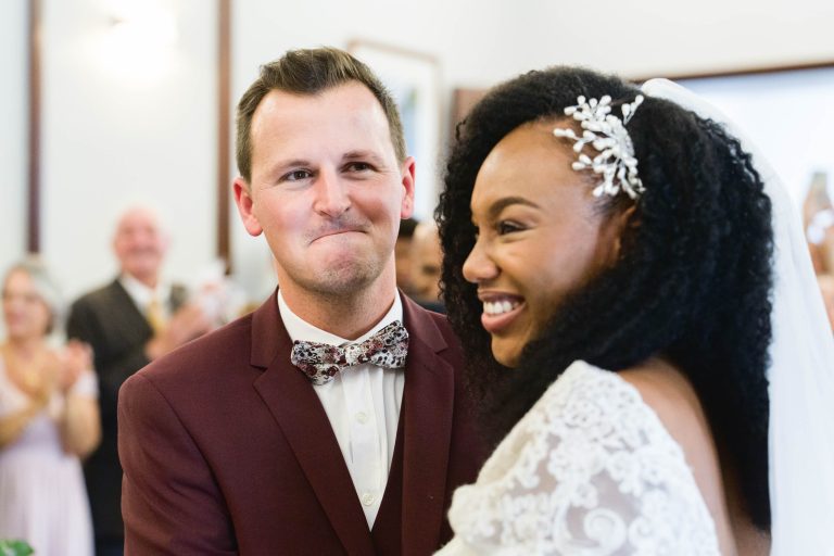 Un marié, vêtu d'un costume marron avec un nœud papillon, se tient debout à côté d'une mariée souriante dans une robe de mariée en dentelle avec un voile lors de leur Mariage Audenge. La mariée a les cheveux bouclés ornés d'un postiche décoratif. Ils sont à l'intérieur avec des spectateurs qui applaudissent en arrière-plan, célébrant l'occasion.