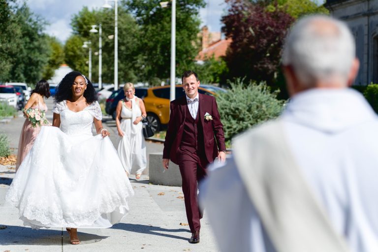 Une mariée en robe blanche et un marié en costume bordeaux marchent sur un chemin extérieur, souriant chaleureusement. La mariée tient sa robe sur le côté. D'autres invités au mariage en tenue élégante marchent derrière eux lors de ce Mariage Audenge. La scène est ensoleillée, avec de la verdure, des voitures garées et des bâtiments en arrière-plan.