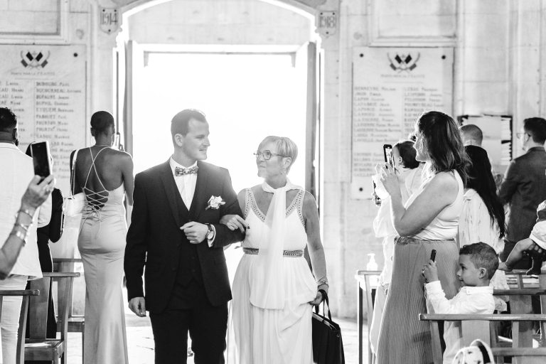 Un marié en costume et nœud papillon marche bras dessus bras dessous avec une femme âgée souriante en robe blanche lors de la cérémonie de mariage de Mariage Audenge. Les invités tiennent des téléphones et prennent des photos. Un jeune enfant au premier rang montre du doigt le couple. L'arrière-plan présente une entrée voûtée ornée de plaques commémoratives.