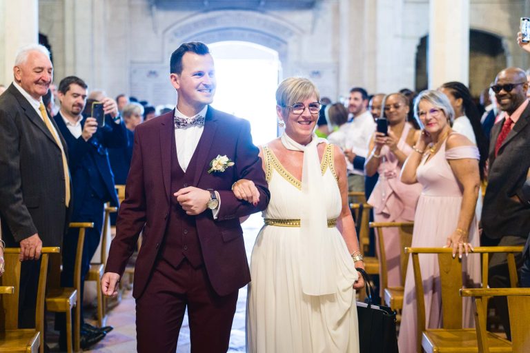 Un cortège de mariage de Mariage Audenge met en scène un homme en costume marron avec un nœud papillon et une boutonnière, marchant bras dessus bras dessous avec une femme en robe blanche aux accents dorés. Ils sourient, se frayent un chemin dans l'allée au milieu d'invités assis et debout qui prennent des photos et applaudissent.