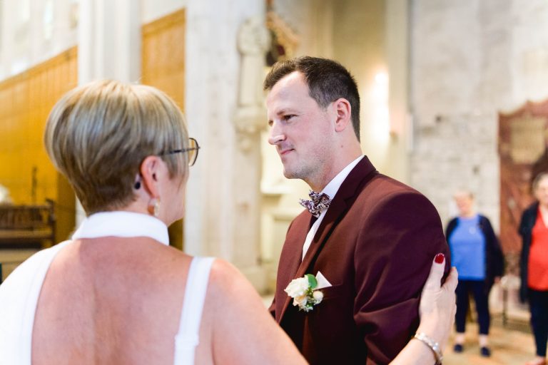 Un homme en costume bordeaux avec une boutonnière fleurie et un nœud papillon à motifs se tient debout à l'intérieur, souriant à une femme aux cheveux blonds courts vêtue d'une robe blanche. Ils se font face, sa main reposant doucement sur son épaule. L'arrière-plan, flou mais animé de personnages et de détails architecturaux, suggère l'occasion joyeuse du Mariage Audenge.