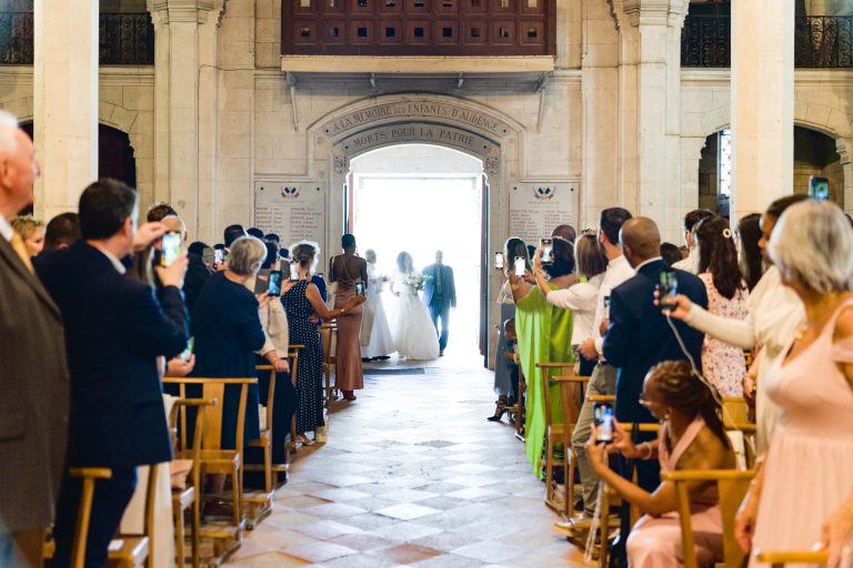 Un couple de mariés, éclairé par la lumière du soleil, marchent dans l'allée d'une grande église décorée remplie d'invités de chaque côté. De nombreux participants sont debout, prennent des photos et certains sont assis. L'église présente de hauts plafonds, des murs en pierre et une entrée aux détails complexes rappelant une cérémonie de mariage Audenge.