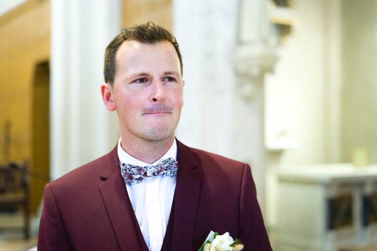 Un homme vêtu d'une veste de costume marron se tient debout dans un décor intérieur, probablement un Mariage Audenge. Il porte une chemise blanche et un nœud papillon à motif floral. Ses cheveux sont soigneusement coiffés et il a un léger sourire sur le visage. L'arrière-plan présente des détails architecturaux flous en blanc.