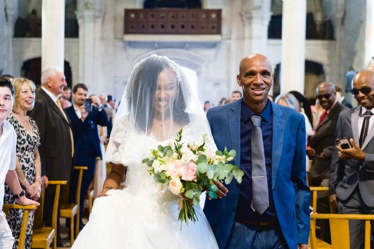 Une mariée souriante, vêtue d'une robe blanche et d'un voile, marche dans l'allée en tenant un bouquet de fleurs, accompagnée d'un homme en costume bleu et cravate. Les invités s'alignent sur les bancs, applaudissent et sourient. L'arrière-plan présente un intérieur grandiose d'une église avec de hautes colonnes et des portes cintrées, ajoutant à l'élégance de ce Mariage Audenge.