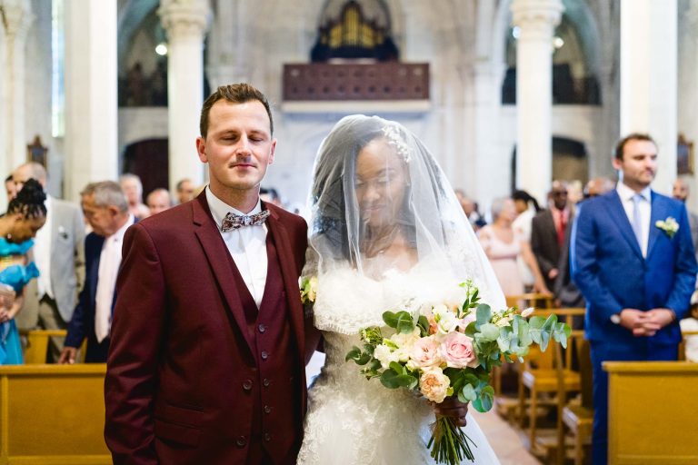 Une mariée et un marié se tiennent côte à côte dans une église lors de leur cérémonie de mariage Audenge. Le marié porte un costume bordeaux avec un nœud papillon, tandis que la mariée est vêtue d'une robe de mariée blanche et d'un voile, tenant un bouquet de fleurs roses et blanches. L'arrière-plan montre les invités du mariage assis sur des bancs en bois, avec des colonnes arquées et un décor d'église.