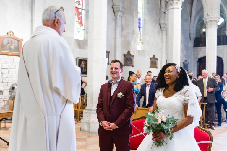 Un couple de mariés se tient devant un prêtre dans une église lors de leur cérémonie de mariage Audenge. Le marié, en costume marron, sourit, tandis que la mariée, en robe de mariée blanche tenant un bouquet, rit. Ils sont entourés d'invités assis sur des bancs, avec des vitraux visibles en arrière-plan.