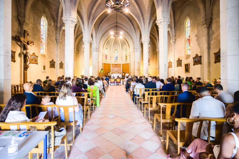 L'image montre l'intérieur d'une église à Audenge, remplie de personnes assises sur des chaises en bois, assistant à une cérémonie de mariage. L'église a de hauts plafonds voûtés, des murs en pierre et des vitraux. À l'avant, un officiant se tient derrière un pupitre, guidant les cérémonies.