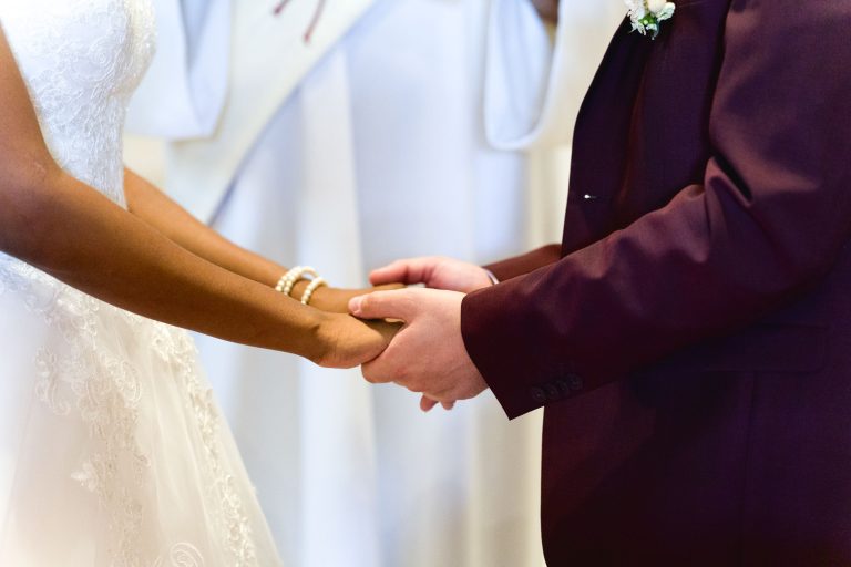 Gros plan d'une mariée et d'un marié se tenant la main lors de leur cérémonie de mariage. La mariée porte une robe en dentelle blanche et un bracelet de perles, et le marié est habillé d'un costume sombre. Un prêtre en robe blanche se tient derrière eux, partiellement visible. La belle scène du mariage d'Audenge transmet l'unité et l'amour.