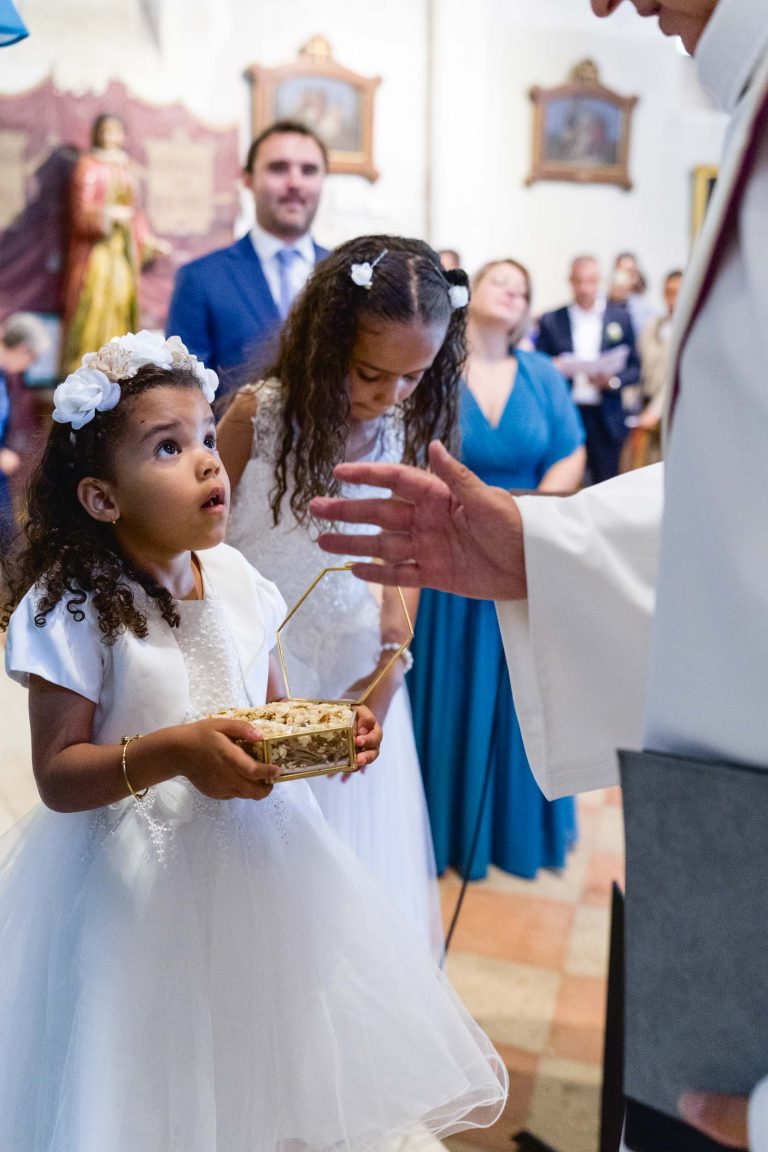 Une jeune fille aux cheveux bouclés et au bandeau de fleurs blanches, vêtue d'une robe blanche, lève les yeux tout en tenant un panier commémoratif en or. Elle se tient dans une église pendant le Mariage Audenge près d'un adulte en robe blanche, avec une autre jeune fille et plusieurs adultes en tenue de soirée en arrière-plan.