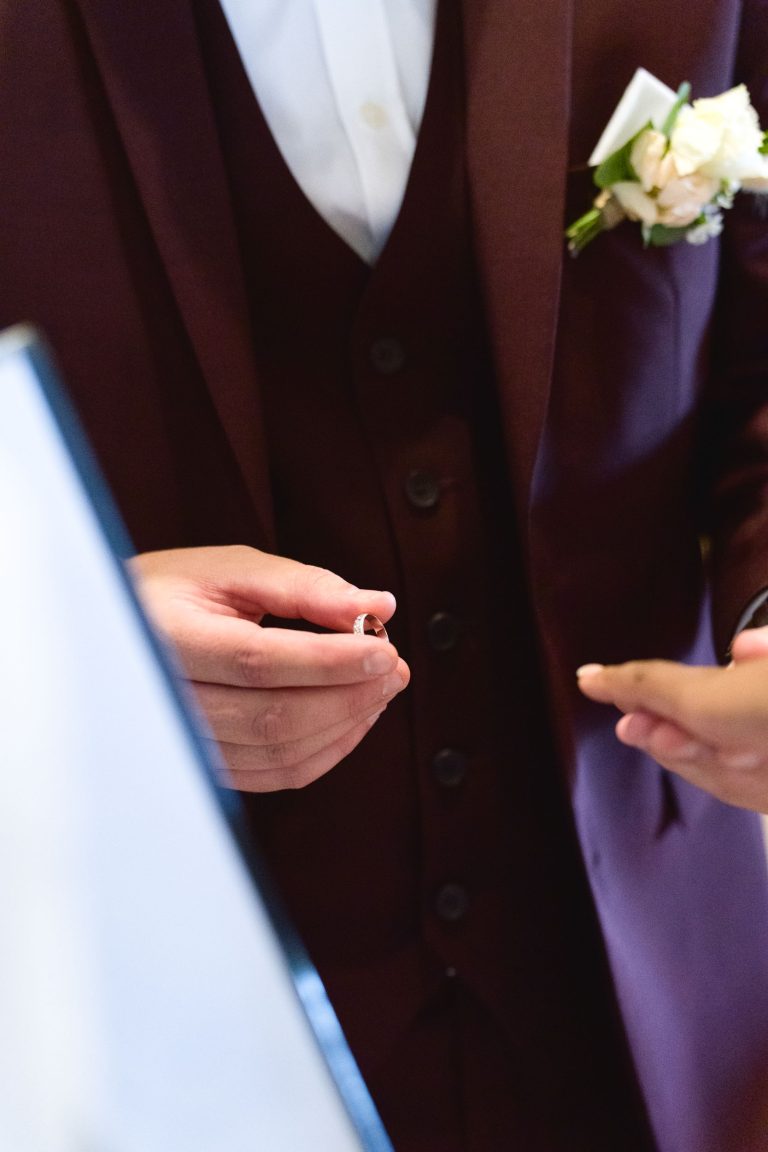 Une personne en costume marron tient une alliance dans ses deux mains. Le costume comprend une chemise blanche, un gilet boutonné et une pochette avec une boutonnière à fleurs. La bague est le point central, la personne semblant être présente au Mariage Audenge, peut-être pendant la cérémonie.