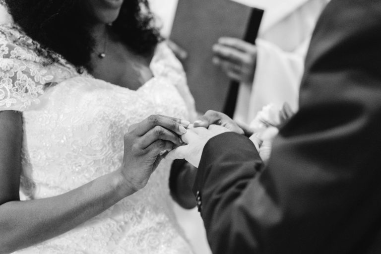 Une photo en noir et blanc capture un gros plan d'une cérémonie de mariage organisée par Mariage Audenge. La mariée, vêtue d'une robe en dentelle complexe, place une bague au doigt du marié. Le marié porte un costume sombre. À l'arrière-plan, une personne tenant un livre se tient debout, probablement en train d'officialiser la cérémonie.