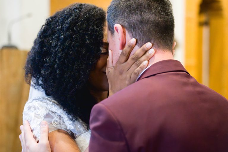 Un couple s'embrasse tendrement lors de leur mariage à Audenge. La mariée, aux cheveux bouclés, porte une robe en dentelle blanche. Le marié, aux cheveux noirs courts, est habillé d'un costume marron. Ils se tiennent doucement le visage et sont entourés d'un décor doux et chaleureux.