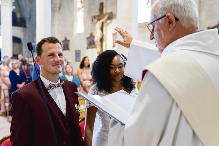 Un couple se tient devant un prêtre lors de leur cérémonie de mariage Audenge dans une église. Le marié porte un costume marron avec un nœud papillon et la mariée est vêtue d'une robe de mariée blanche. Le prêtre, vêtu de vêtements liturgiques blancs, tient un livre et lève la main en signe de bénédiction. Les invités regardent en arrière-plan.