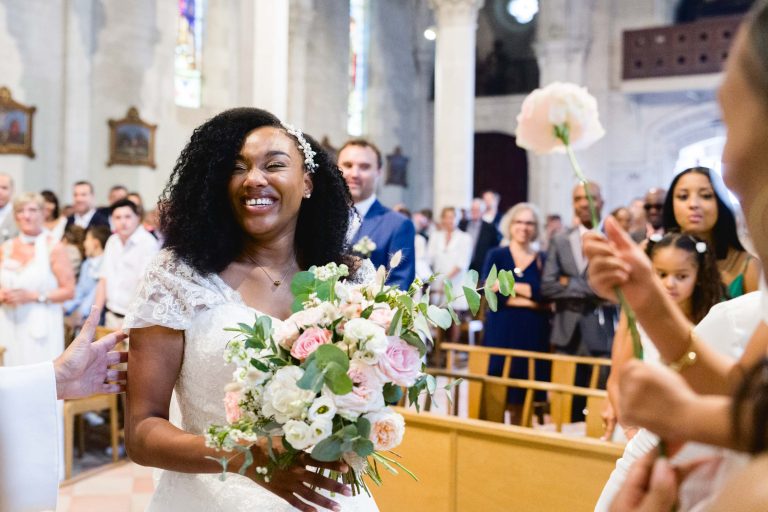 Une mariée joyeuse aux cheveux noirs bouclés et une fleur dans les cheveux, vêtue d'une robe de mariée en dentelle blanche, tient un bouquet de fleurs pastel tout en marchant dans l'allée à l'intérieur de l'église de Mariage Audenge. Les invités, vêtus de tenues semi-formelles, regardent avec le sourire les vitraux et les colonnes qui ajoutent à la beauté du décor.