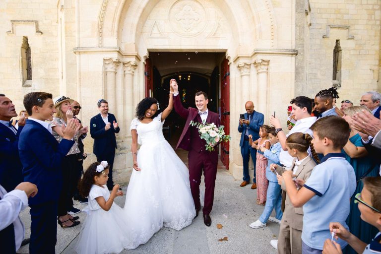 Une mariée souriante en robe blanche et un marié en costume bordeaux marchent main dans la main depuis l'église historique du Mariage Audenge. La mariée tient un bouquet et leurs mains sont levées. Les invités, dont des enfants, applaudissent et jettent des confettis. L'église en pierre a de grandes portes cintrées et l'ambiance est joyeuse et festive.