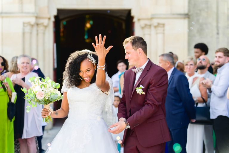 Une mariée montre joyeusement son alliance tout en tenant un bouquet dans son autre main. Elle porte une robe de mariée blanche et se tient à côté de son marié souriant, qui est habillé d'un costume marron avec un nœud papillon. Ils sont entourés d'amis et de membres de la famille célébrant leur mariage Audenge à l'extérieur d'un bâtiment historique.