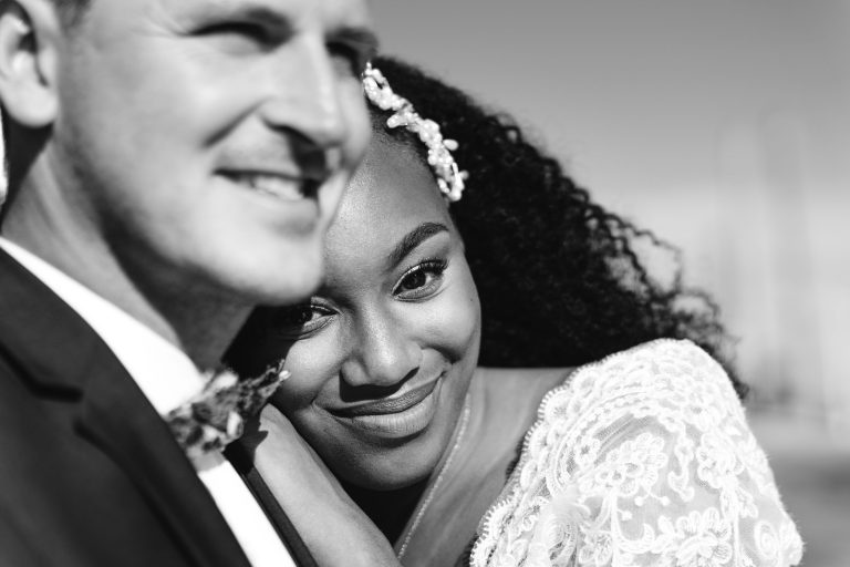 Une photo en noir et blanc d'un couple de jeunes mariés lors de leur mariage Audenge. Le marié, en costume, sourit au premier plan tandis que la mariée, parée d'un accessoire de cheveux fleuri et d'une robe en dentelle, pose sa tête sur son épaule. Tous deux rayonnent de contentement tandis que l'arrière-plan se brouille doucement.