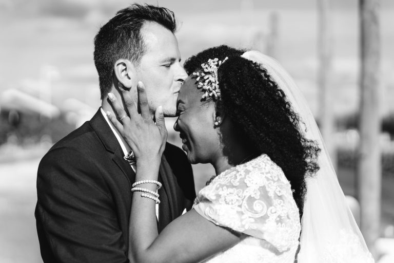 Une photo en noir et blanc capture un moment de joie entre un couple de jeunes mariés lors de leur Mariage Audenge. Le marié, en costume, embrasse le front de la mariée, qui porte une robe de mariée en dentelle et un voile. Elle sourit largement, sa main reposant doucement sur son cou. La scène se déroule en extérieur avec un arrière-plan flou.