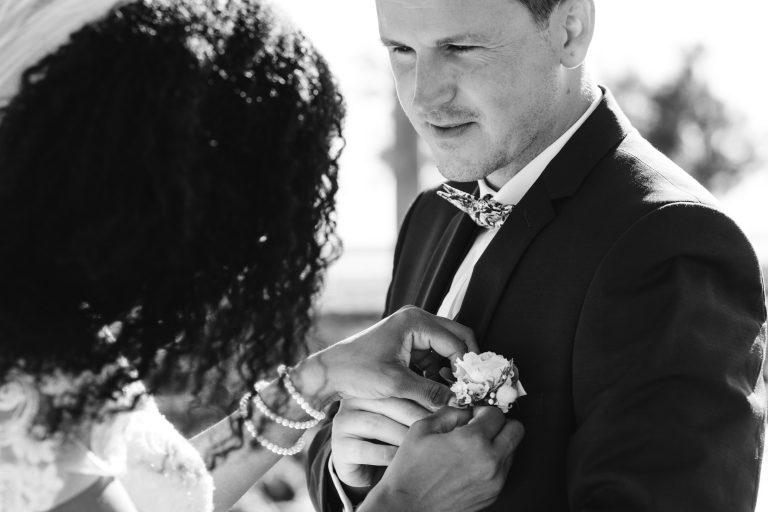 Une femme en robe de mariée aide à ajuster une boutonnière de fleurs sur le revers d'un costume d'homme. L'homme regarde la boutonnière avec un doux sourire, incarnant l'essence de Mariage Audenge. L'arrière-plan est flou, avec la lumière vive du soleil projetant des ombres et ajoutant un effet de rêve à la scène.