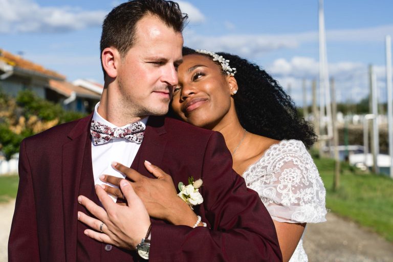 Un marié vêtu d'un costume bordeaux avec un nœud papillon fleuri regarde sur le côté tandis qu'une mariée en robe de dentelle blanche avec un postiche fleuri, l'enlaçant par derrière, pose sa main sur sa poitrine. Ils sont à Mariage Audenge, avec le ciel bleu et les mâts de la marina en arrière-plan. Tous deux ont des expressions sereines et satisfaites.