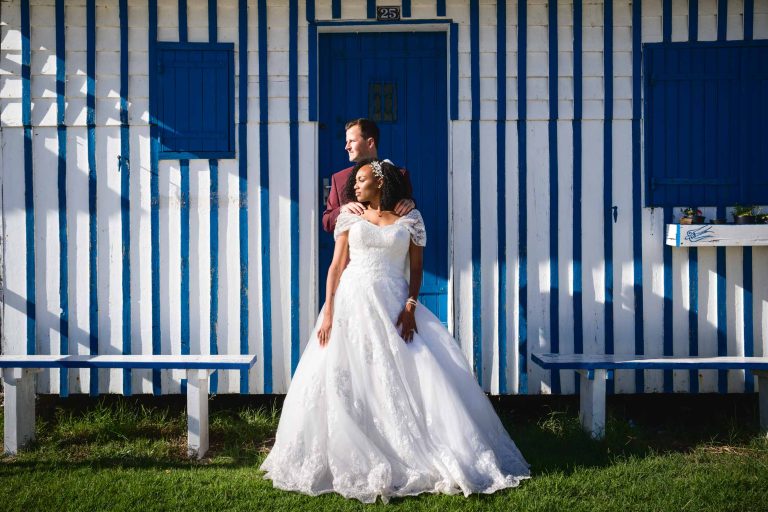 Une mariée en robe blanche et un marié en costume sombre se tiennent devant le charmant bâtiment rayé bleu et blanc, capturant l'essence d'un Mariage Audenge pittoresque. La mariée regarde vers l'avant tandis que le marié enroule affectueusement ses bras autour de sa taille, tandis que la lumière du soleil joue sur la scène.