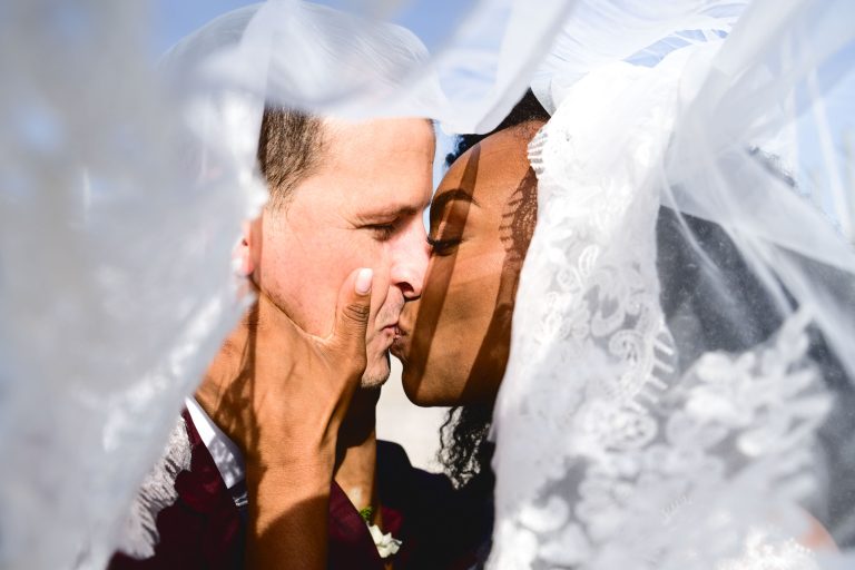 Gros plan d'un couple de jeunes mariés s'embrassant sous un voile blanc translucide lors de leur Mariage Audenge. Les mains du marié tiennent doucement le visage de la mariée tandis que ses mains reposent sur son cou. Le fond est un ciel bleu vif, et la douce lumière filtre à travers la dentelle du voile, créant une atmosphère intime et romantique.