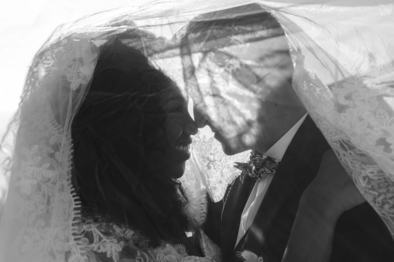 Photo en noir et blanc d'un couple le jour de leur mariage à Mariage Audenge. Ils sont enlacés sous un voile de dentelle, leurs fronts se touchant et souriant. La lumière du soleil projette de douces ombres à travers le voile, renforçant l'atmosphère romantique. Le marié porte un costume et la mariée a les cheveux bouclés.