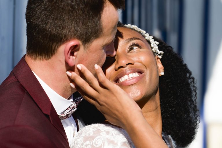 Dans leur Mariage Audenge, les mariés se regardent amoureusement, de près. Elle sourit largement, lui touche la joue de la main. Le marié, vêtu d'un costume marron, la regarde de travers. La mariée porte une robe blanche, les cheveux ornés d'un postiche. Des lignes verticales bleues sont floues à l'arrière-plan.