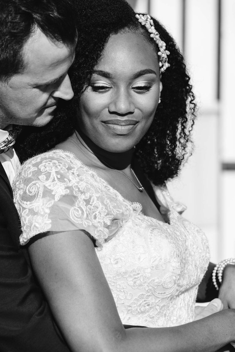 Une photo en noir et blanc d'un couple. La mariée, souriant doucement, porte une robe de mariée en dentelle aux détails floraux et un collier de perles au poignet. Elle a les cheveux bouclés ornés d'un postiche. Le marié, vêtu d'un costume, l'embrasse doucement par derrière, la regardant avec amour, un souvenir parfait du Mariage Audenge.