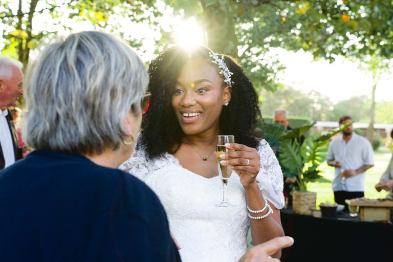 Une mariée en robe blanche tient un verre de champagne et sourit tout en discutant avec une femme plus âgée portant des lunettes au Mariage Audenge. La lumière du soleil filtre à travers les arbres, illuminant ses cheveux décorés d'un couvre-chef fleuri. D'autres personnes sont visibles à l'arrière-plan, profitant de la verdure.