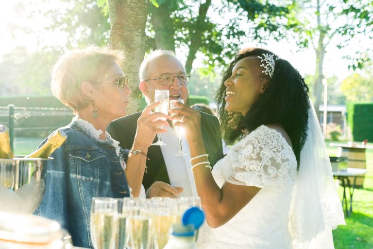 Une mariée en robe blanche et voile, tenant une flûte de champagne, sourit et porte un toast avec un homme âgé en costume et une femme âgée portant des lunettes de soleil et une veste en jean. Ils sont à l'extérieur, entourés d'arbres et de soleil lors de ce qui semble être une belle célébration du Mariage Audenge, près d'une table remplie de plusieurs coupes de champagne.