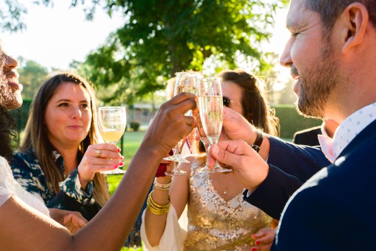 Un groupe de personnes se rassemble à l'extérieur lors d'un mariage Audenge, levant leurs verres pour porter un toast. Ils sont habillés de manière formelle, l'un portant une robe à paillettes et les autres des costumes et des tenues élégantes. Le fond vert et la lumière du soleil filtrant à travers les arbres créent une atmosphère festive et joyeuse.